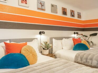 photo of a 70s/80s retro themed hotel room with three stripes (one orange, one red, one black) painted along the white walls and vinyl record covers hanging above the stripes as decoration. the two queen beds feature white cotton linens and gray fur throws, as well as three primary color (red, yellow, blue) accent pillows on each bed. the bedside tables have small black metal lights above them and a plant on them.