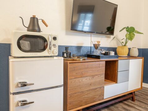 a photo featuring a room in a boutique luxury motel in bandon, oregon showcasing some of the room amenities, including a small white mini-fridge, a white microwave on top, a black tea kettle, a mid-century modern media cabinet with eclectic home decor and a small plant on top. the wall is bordered by a blue stripe that spans the bottom half of the wall, and has a large TV.