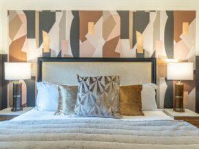 photo of a luxury oregon motel bed in 100% white cotton linens, isotonic pillows, a gray vegan fur throw, and silver and gold accent pillows, with a tan headboard with a dark wood border. there is an abstract wallpaper behind the headboard with neutral-colored organic shapes and two nightstands with matching lamps that have a black base and white lampshade.