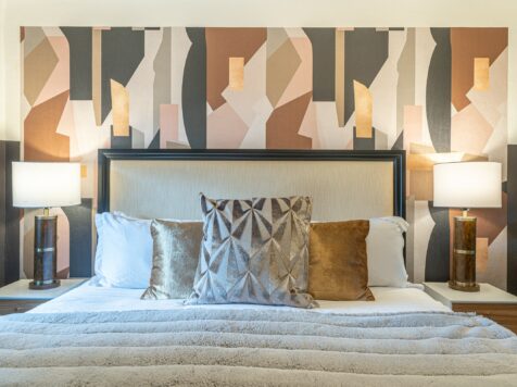 photo of a luxury oregon motel bed in 100% white cotton linens, isotonic pillows, a gray vegan fur throw, and silver and gold accent pillows, with a tan headboard with a dark wood border. there is an abstract wallpaper behind the headboard with neutral-colored organic shapes and two nightstands with matching lamps that have a black base and white lampshade.