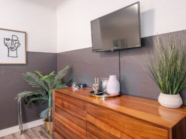 a corner of a motel room in a dog-friendly motel on the oregon coast featuring a midcentury modern media cabinet with two plants in white planters, eclectic home decor including a gold hand making a piece sign and an empty white vase, and a smart tv hanging above the cabinet.