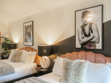 photo of two queen beds in a southern oregon coast motel with light pink headboards, white linens, gray vegan fur throws, and gray and white accent pillows. the wall is painted with a diagonal black portion cutting across the wall halfway and features two black and white photos behind the bed, one of whitney houston and one of madonna. the third is a poster of cindy crawford. the room has two nightstands with unique modern sidetable lamps with white bases and black lampshades, as well as a fiddleleaf plant.