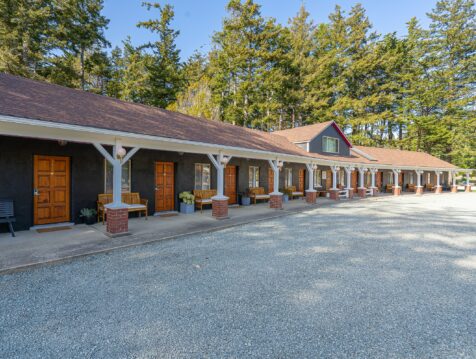 photo of an oregon coastal motel in bandon painted black with brown wooden doors, a brown roof, white pillars with brick bottoms, and the edge of a packed gravel parking lot