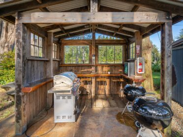 photo features a shared grill area at a bandon oregon rv park. the grill area has a covered wooden structure with a counter and wooden and metal barstools, a nexgrill, and two black weber grills.
