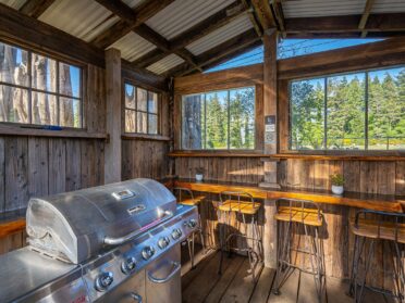 photo features a shared grill area at a bandon oregon rv park. the grill area has a covered wooden structure with a counter and wooden and metal barstools and a silver nexgrill.