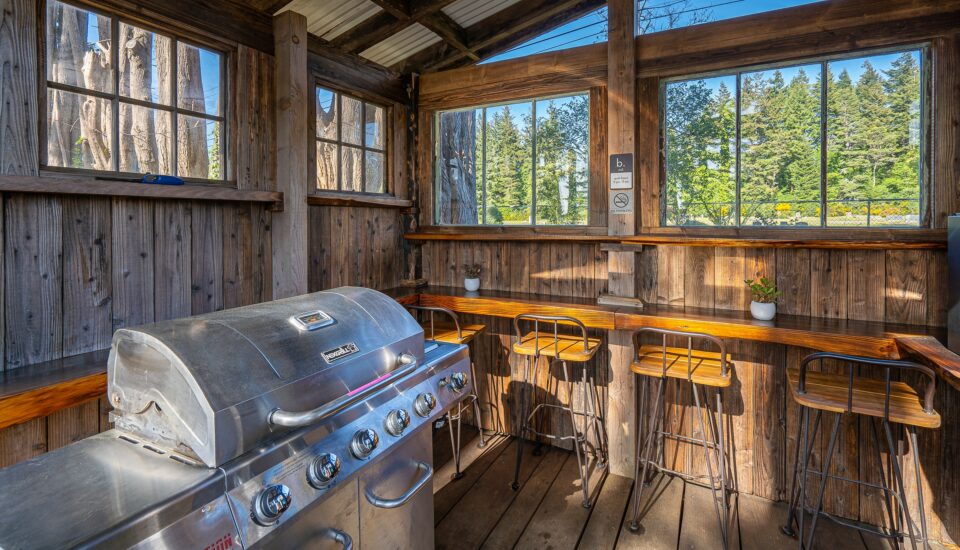 photo features a shared grill area at a bandon oregon rv park. the grill area has a covered wooden structure with a counter and wooden and metal barstools and a silver nexgrill.