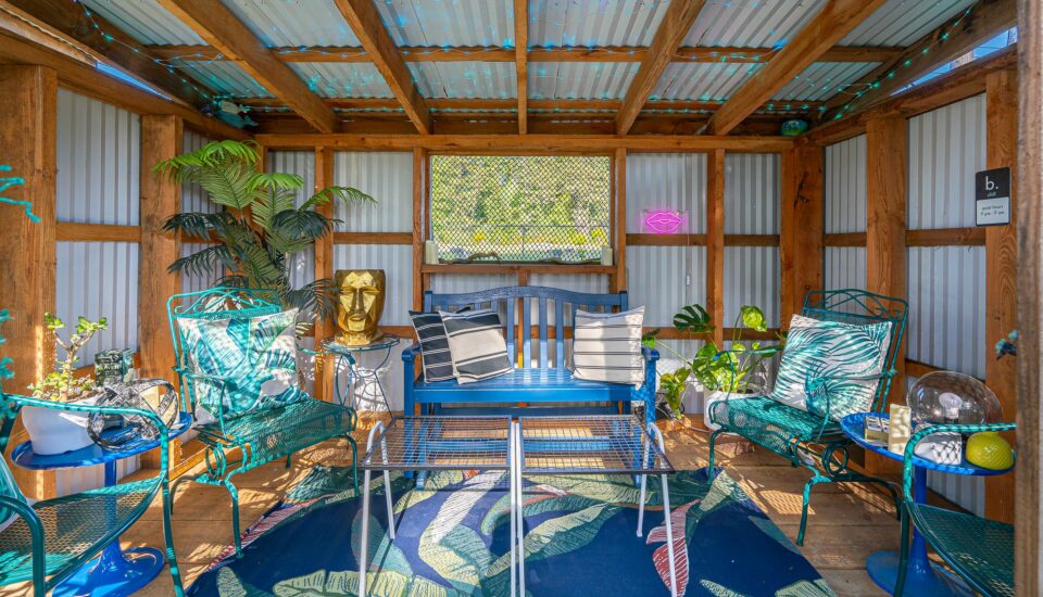 photo features a cannabis hut at a motel in bandon oregon. cannabis hut is an outdoor covered seating area with eclectic decor, including a blue rug with bright green leaves, blue and green chairs and a bench, a table with tic-tac-toe, and a gold planter that looks like a head, as well as a neon lips sign.