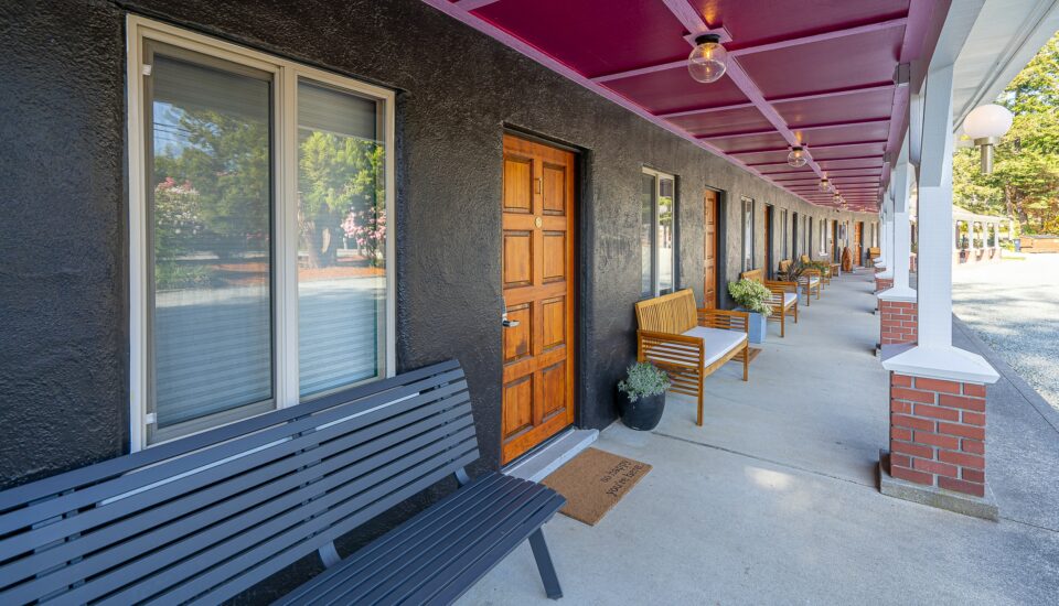 photo of an outdoor thoroughfare at a boutique motel and rv park in bandon, oregon. the exterior walls are painted black, with a pink awning and brown wooden doors. the pathway is cement and has planters filled with colorful flowers, and black and rattan benches with white cushions.