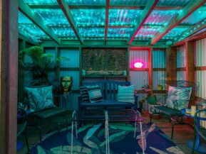 photo features a cannabis hut at a bandon oregon hotel with shared outdoor spaces. cannabis hut is an outdoor covered seating area with eclectic decor, including a blue rug with bright green leaves, blue and green chairs and a bench, a table with tic-tac-toe, and a gold planter that looks like a head, as well as a neon lips sign. the hut is lit with ambient green light for a fun, moody atmosphere.