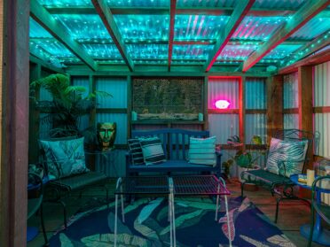 photo features a cannabis hut at a bandon oregon hotel with shared outdoor spaces. cannabis hut is an outdoor covered seating area with eclectic decor, including a blue rug with bright green leaves, blue and green chairs and a bench, a table with tic-tac-toe, and a gold planter that looks like a head, as well as a neon lips sign. the hut is lit with ambient green light for a fun, moody atmosphere.