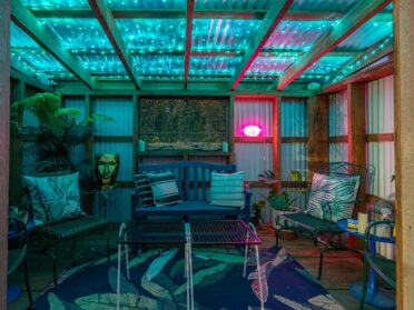 photo features a cannabis hut at a bandon oregon hotel with shared outdoor spaces. cannabis hut is an outdoor covered seating area with eclectic decor, including a blue rug with bright green leaves, blue and green chairs and a bench, a table with tic-tac-toe, and a gold planter that looks like a head, as well as a neon lips sign. the hut is lit with ambient green light for a fun, moody atmosphere.