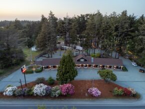 a drone shot looking straight ahead at a one-story motel in bandon oregon. the shot features a full picture of the oregon coast hotel and bandon rv park, which includes a black building with a brown roof and warm white lights along white pillars lining the outdoor walkway, bright flowers and foliage lining the edge of the packed gravel parking lot, and a variety of tall trees scattered throughout and around the campsites and rv park behind the motel.