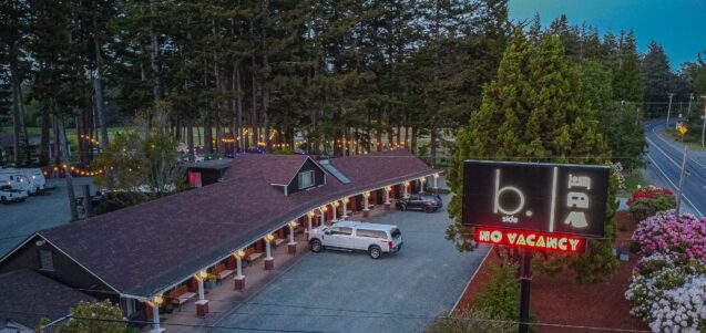 a drone shot of a one-story dog-friendly hotel in bandon oregon. the shot features a full picture of the oregon coast hotel and bandon rv park, which includes a black building with a brown roof and warm white lights along white pillars lining the outdoor walkway, bright flowers and foliage lining the edge of the packed gravel parking lot, and a variety of tall trees scattered throughout and around the campsites and rv park behind the motel.