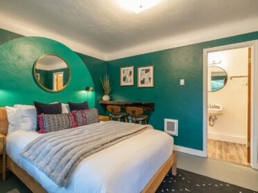 photo of a coos bay hotel room with dark green paint and a lighter green arch behind the warm wood midcentury modern bed frame. the bed has white linens, a gray vegan fur throw blanket, and blue and purple and black accent pillows. a small round mirror sits above the headboard of the bed, and a midcentury modern nightstands in the same warm wood as the bed sits to the left under a black metal light hanging from the wall. the room also has a small wood desk attached to the far right wall, with two modern barstools, and shows an open door to the bathroom featuring a round mirror and white sink.